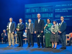 Na scenie stoi siedem osób ustawionych w rzędzie. Każda trzyma białą różę. W środku stoi wysoki mężczyzna w ciemnym garniturze. Obok niego stoi kobieta z ciemnymi włosami w zielonej bluzce i zielonej spódnicy. Pozostałe osoby mają garnitury lub eleganckie stroje. W tle siedzą uczestnicy wydarzenia. Na ekranie po prawej stronie widoczne są napisy: osoby z niepełnosprawnością intelektualną, II miejsce: Patrycja Jarychowicz i Szymon oraz czyta Małgorzata Zawadzka. Część tekstu z nazwiskami jest poza kadrem i pozostaje nieczytelna.