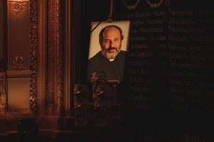 Portret księdza stoi na drewnianej sztaludze. Mężczyzna ma brodę, ciemne ubranie i koloratkę. Patrzy prosto przed siebie. Pod portretem stoją maski teatralne ustawione jedna na drugiej. Obok widać zdobioną ścianę ze złotymi elementami. Tło jest ciemne.