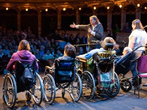 Scena teatralna podczas występu ujęta od kulis. Na pierwszym planie, tyłem do widza, ale przodem do widowni siedzą cztery osoby na wózkach inwalidzkich. Po lewej kobieta w fioletowej bluzie z kapturem, z ciemnymi włosami, siedzi na czarnym wózku. Obok niej kobieta w czarnej bluzie, z krótkimi ciemnymi włosami, na niebiesko-czarnym wózku. Po prawej dwie osoby, jedna z nich leży częściowo w kolorowym kocu na wózku. Na scenie po prawej stronie stoją trzy osoby. Mężczyzna pośrodku sceny ma jasne włosy, długą koszulę i wyciąga prawą rękę w kierunku publiczności, mówiąc do mikrofonu. Po prawej stoją kobieta w białej bluzce oraz mężczyzna w kamizelce, oboje trzymają mikrofony. Za nimi stoi fioletowe krzesło. Za sceną widoczna jest duża publiczność, rzędy krzeseł są wypełnione. Sufit sali zdobią rzędy małych lamp.