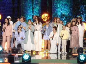 Na scenie stoi grupa osób trzymających mikrofony. Część z nich śpiewa, inni patrzą w różne strony. Uczestnicy mają różne stroje w jasnych i pastelowych kolorach — płaszcze, koszule, spódnice i spodnie. W tle widać dekoracje w zielonych barwach i okrągłe, pomarańczowe światła sceniczne. Podłoga sceny odbija światło reflektorów.