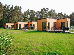 Pięć identycznych, parterowych domków stoi w rzędzie na tle lasu sosnowego. Domki mają jasne, drewniane elewacje i czarne obramowania dużych okien oraz drzwi tarasowych. Przed każdym domkiem jest drewniany taras z czarnymi, metalowymi balustradami, wyposażony w stoliki i krzesła. Z przodu rozciąga się duży, zielony trawnik. Przy tarasach widać łagodnie nachylone, drewniane podjazdy z balustradami, dostosowane dla osób z niepełnosprawnościami. W lewym dolnym rogu znajduje się fragment krzewu z białymi kwiatami.