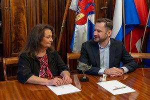 Kobieta i mężczyzna patrzą na siebie i siedzą przy stole. Kobieta ma długie, ciemne włosy. Ubrana jest w czarny żakiet i kwiecistą bluzkę. Ręce trzyma na blacie stołu na białej teczce. Mężczyzna siedzi po jej prawej stronie. Ma krótkie ciemne włosy. Czarną marynarkę i białą koszulę. Przed nim na stole leży biała teczka. Na stole stoi również statuetka ptaszka. W tle boazeria i biało-niebiesko-czerwone flagi, w tym flaga z herbem miasta Krakowa.