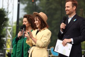 Trzy osoby stoja na scenie plenerowej. Trzymają mikrofony. Kobieta po lewej ma na sobie zielone ubranie. Pośrodku stoi osoba w jasnym płaszczu i kapeluszu. Po prawej osoba w ciemnym garniturze trzyma teczkę. W tle widoczne są drzewa i elementy scenicznego oświetlenia.