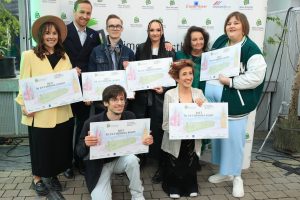 Osiem osób stoi w grupie w dwóch rzędach. Uśmiechają się. Ubrani są elegancko. W środku grupy stoi kobieta w zielonej tunice, która obejmuje młodego chłopca. W tylnym rzędzie stoi mężczyzna w garniturze. Pozostała szóstka to młodzi wokaliści trzymający w rękach bilety do Zaczarowanej Krainy. Za ich plecami ścianka do fotografii z logo fundacji Anny Dymnej „Mimo Wszystko”. Po prawej stronie pień drzewa w donicy oraz biała siatka ogrodzenia.