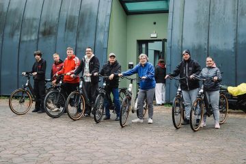 Grupa ośmiu osób stoi z rowerami elektrycznymi przed wejściem do budynku. Pozują do zdjęcia i uśmiechają się. Ubrani są w kurki i niektórzy w czapki.