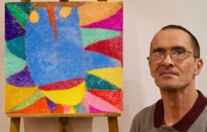Jasne pomieszczenie. Mężczyzna stoi obok kolorowego, abstrakcyjnego obrazu. Dzieło jest umieszczone na sztaludze. Mężczyznę widać od ramion w górę. Ma na sobie okulary i ubrany jest w koszulkę z kołnierzykiem. Obraz na sztaludze składa się z intensywnych, kolorowych i nieregularnych kształtów.