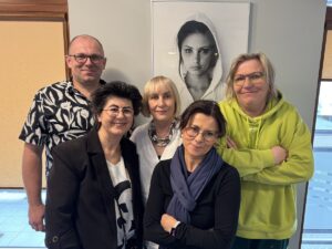Mężczyzna w okularach i cztery kobiety stoją na tle zdjęcia Anny Dymnej. Uśmiechają się. Mężczyzna ma na sobie biało-granatową koszulę z krótkim rękawem. Dwie kobiety ubrane są w ciemne kolory. Jedna ma na sobie białą bluzkę i kolorowe korale. Druga ubrana jest w luźną, żółtą bluze.