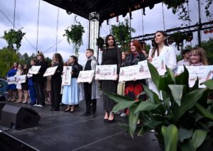 Grupa osób stoi w rzędzie na scenie plenerowej. Trzymają duże dyplomy w obu rękach. Osoby noszą eleganckie stroje. Nad sceną wiszą zielone rośliny i lampy sceniczne. Na pierwszym planie widać duże, zielone liście częściowo zasłaniające widok. W tle znajdują się drzewa oraz konstrukcja sceny.